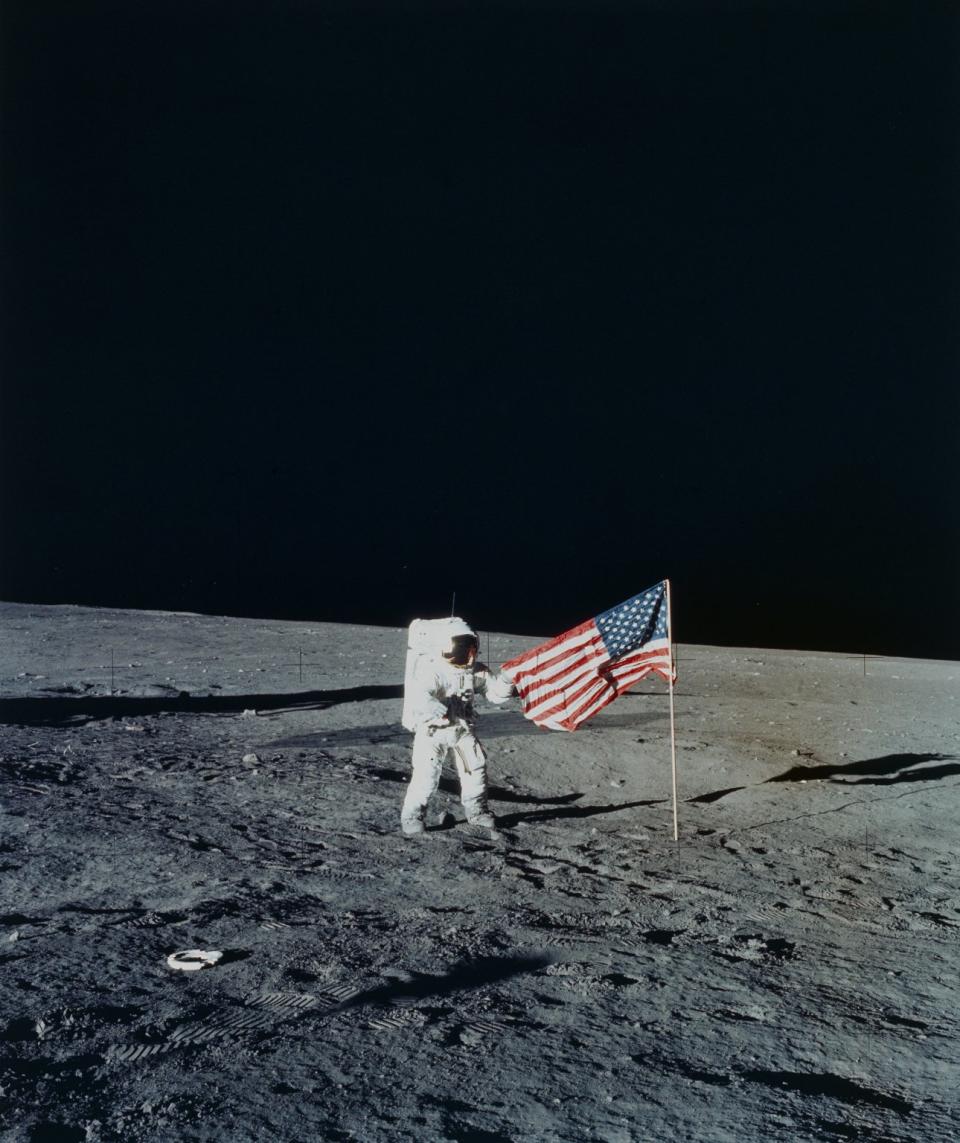 an astronaut in a white space suit spreads an American flag over a dusty blue moon