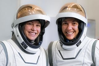 Two women wearing white space suits are seen from the chest up.  Their copper colored visors are lifted up to reveal their faces hidden within their helmets.  Those in the white room are smiling.