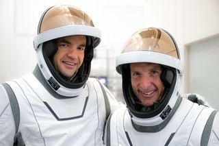 Two men in white space suits are seen from the chest up.  Their copper colored visors are lifted up to show their faces hidden inside their helmets.  Those in the white room are smiling.