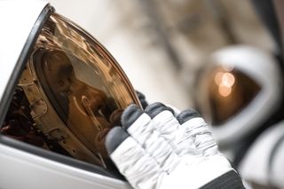 A close-up of an astronaut's helmet with a white shell from the left, to the center of the image.  A copper-shaded visor is straight across the dark inner face.  White gloves with a black tip test over the visor.