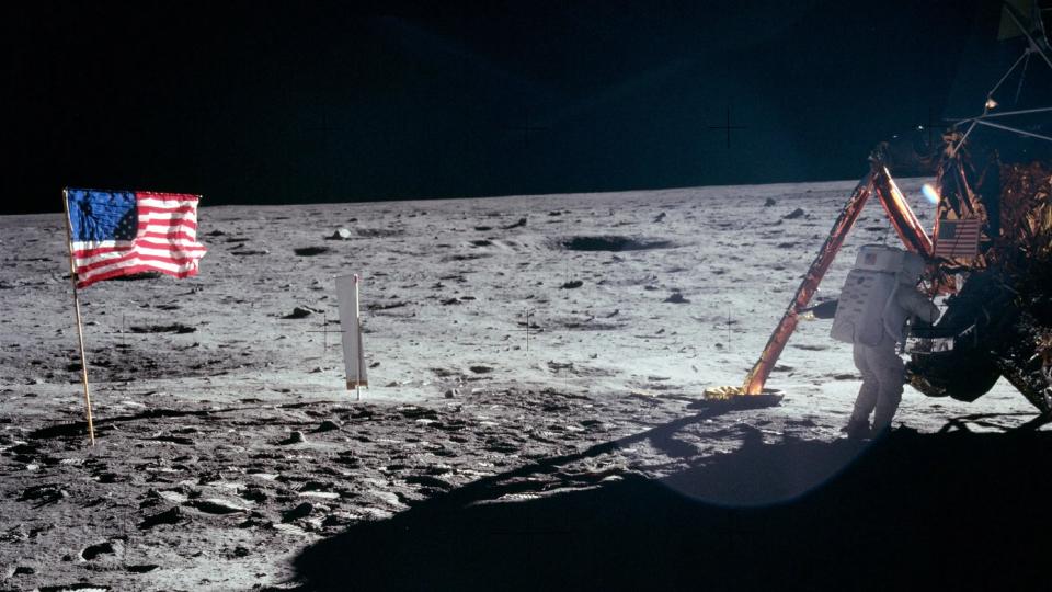 An astronaut in a space suit with a golden face shield walks on a dusty and cloudy moon.